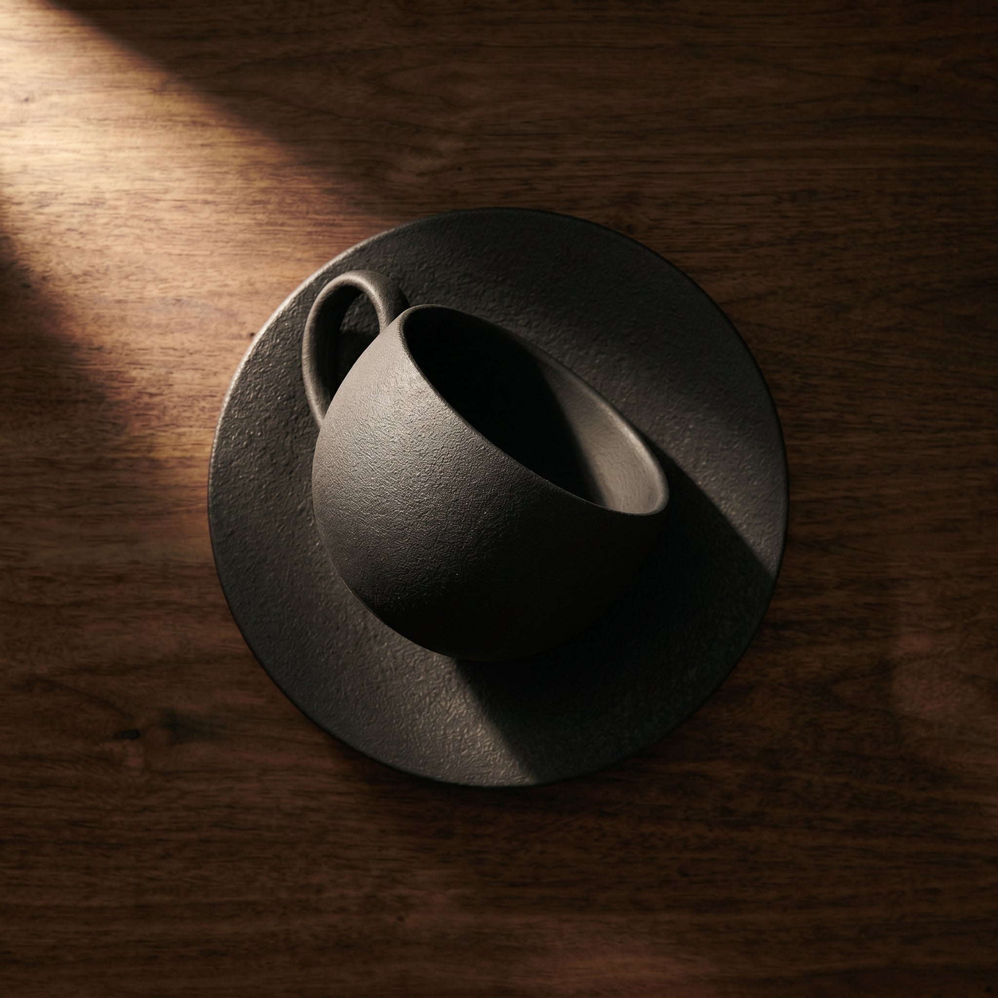 High-angle top-down perspective of the empty dark gray Basalt cup and saucer, showcasing the unpolished granular stone texture under sharp directional window light.