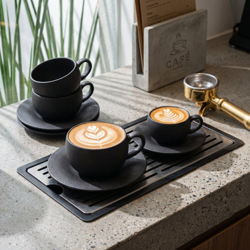 A collection of coarse, volcanic-textured Ando espresso and latte cups in Basalt black arranged across a stone café countertop next to a commercial portafilter.