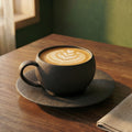 The Basalt variant of The Ando textured ceramic cup and saucer displaying detailed poured latte art on a rich walnut tabletop.