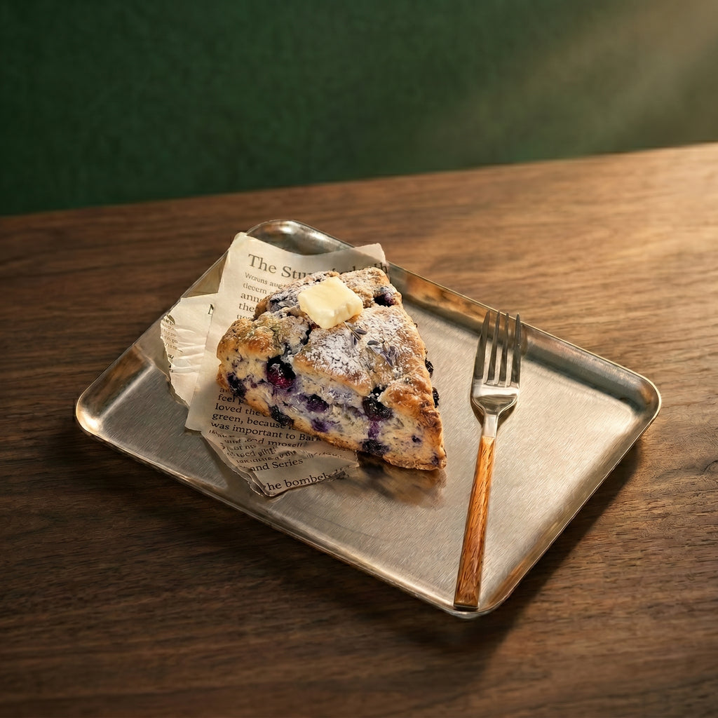 A gourmet blueberry scone with butter resting on parchment liner atop The Carson brushed service platter with a wood-handled fork on a walnut table.
