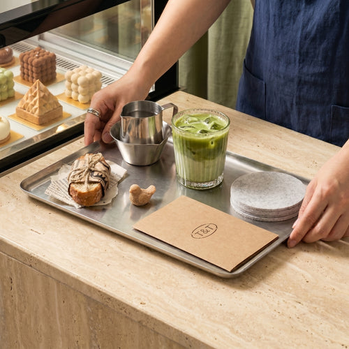 A café barista holding a large Carson brushed metal serving platter arranged with an iced matcha latte, a pastry, ceramic saucers, and a stainless steel pitcher.