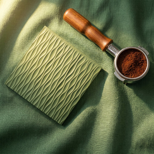 The Conrad contour silicone tamping mat in laurel green alongside a wooden handle portafilter filled with espresso grounds on a textured green linen fabric.