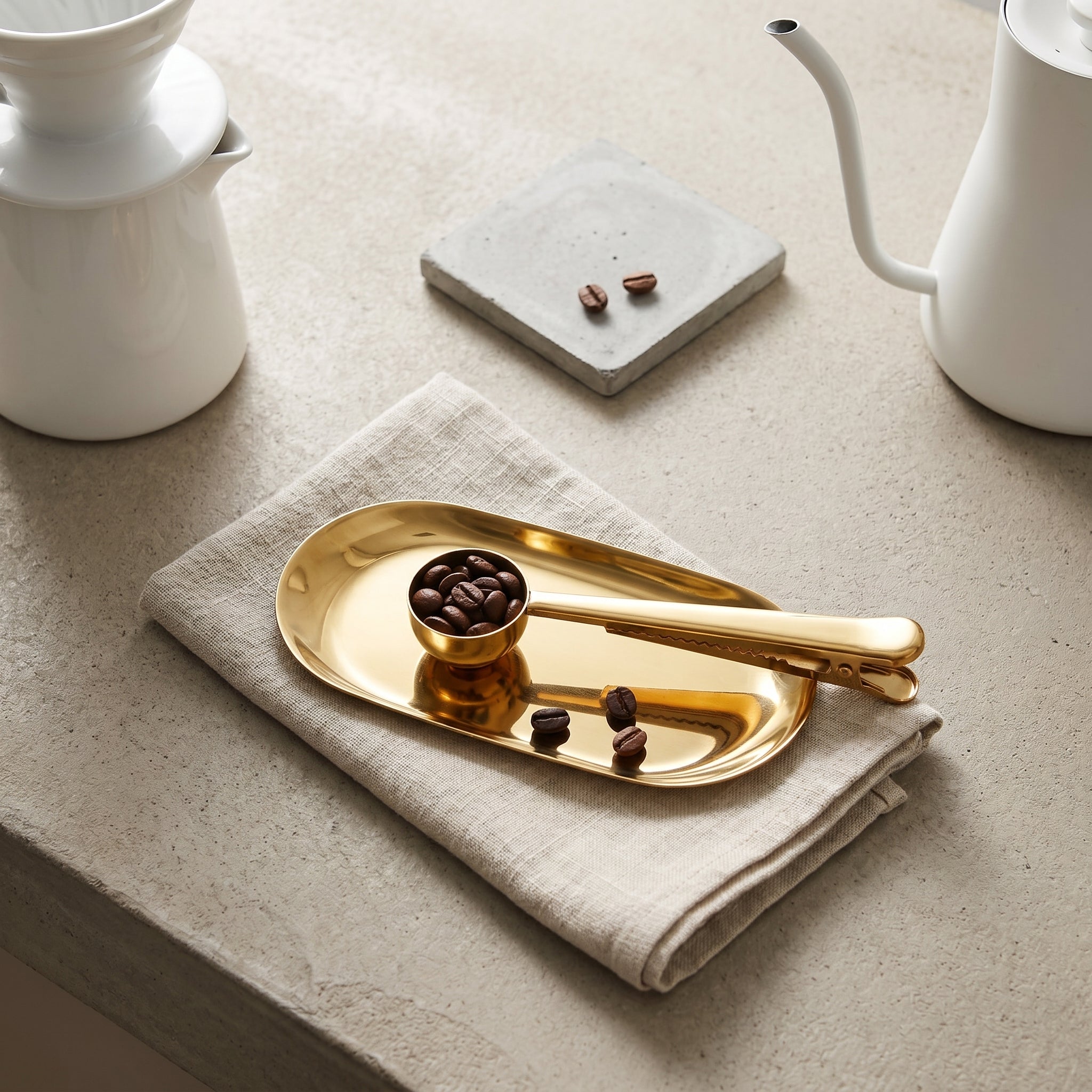 High-angle shot of the gold Dalton coffee measure resting on a linen napkin inside a polished metal utility tray next to loose coffee beans.