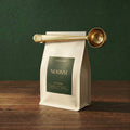 The Dalton tapered dosing clamp in satin brass sealing a beige single-origin coffee bag horizontally on a rich walnut surface.