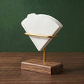 Architectural three-quarter view of the Gable floating filter cradle in solid walnut and brass holding white cone paper filters on a dark wood table against a forest green background.