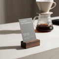 The Radcliffe walnut menu block holding a vertical grey informational card next to a clear glass pour-over coffee carafe on a stone brew bar workstation.