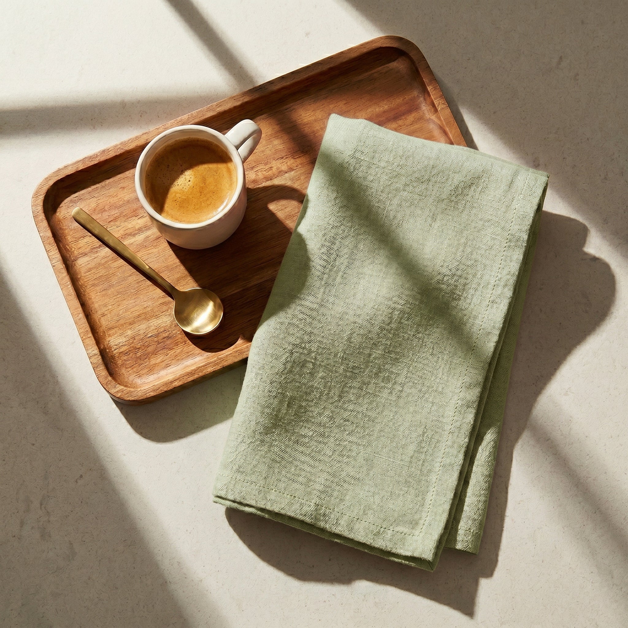 The Sutton table linen napkin in sage green displayed on a wooden service tray next to a ceramic espresso cup.