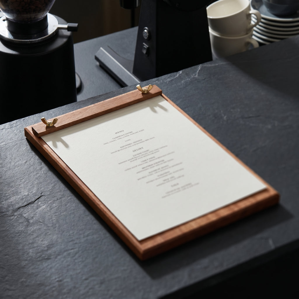 The large-format Plenary Deck Talbot coffee shop menu holder featuring a full beverage registry resting on a textured black stone workspace under dramatic lighting.