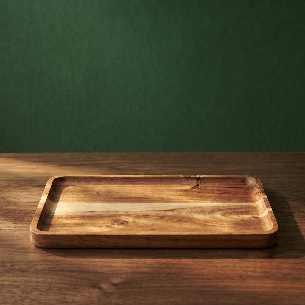 Single empty Vance beveled rectangular acacia wood presentation tray on a dark timber surface against a deep forest green wall under hard direct sunlight.