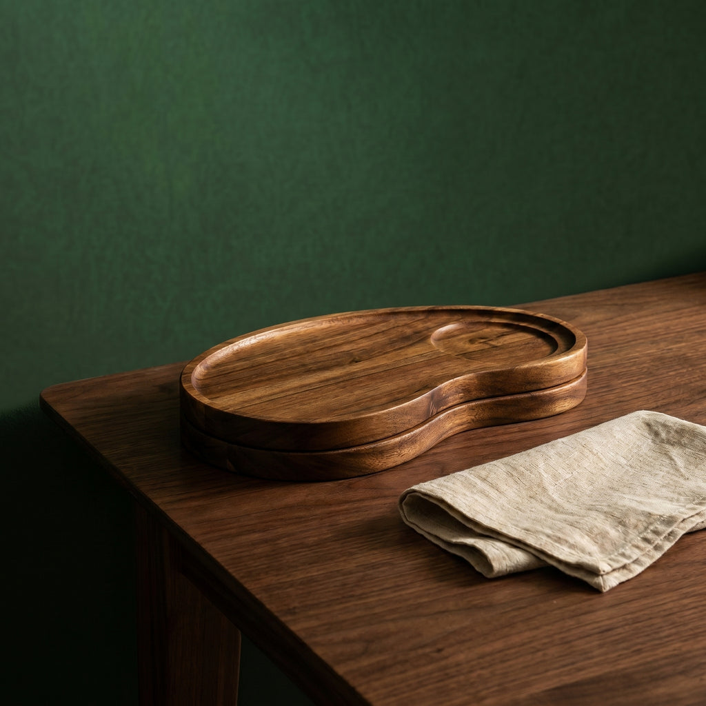 Two pieces of the Winston asymmetric cashew wood trays stacked neatly together next to a stonewashed linen napkin on a counter.